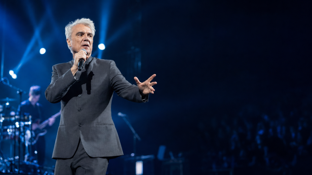 デヴィッド・バーンが青と白の照明に包まれたステージでマイクを手に歌う、2026年来日公演をイメージした実写風ビジュアル